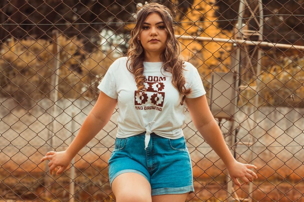 Confident woman in denim shorts and white shirt poses by a chain link fence in warm daylight.