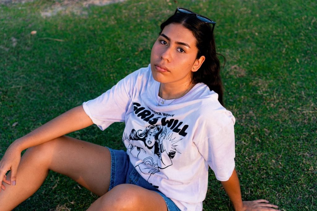 A young woman sits on grass wearing a graphic tee, enjoying a relaxed outdoor moment.