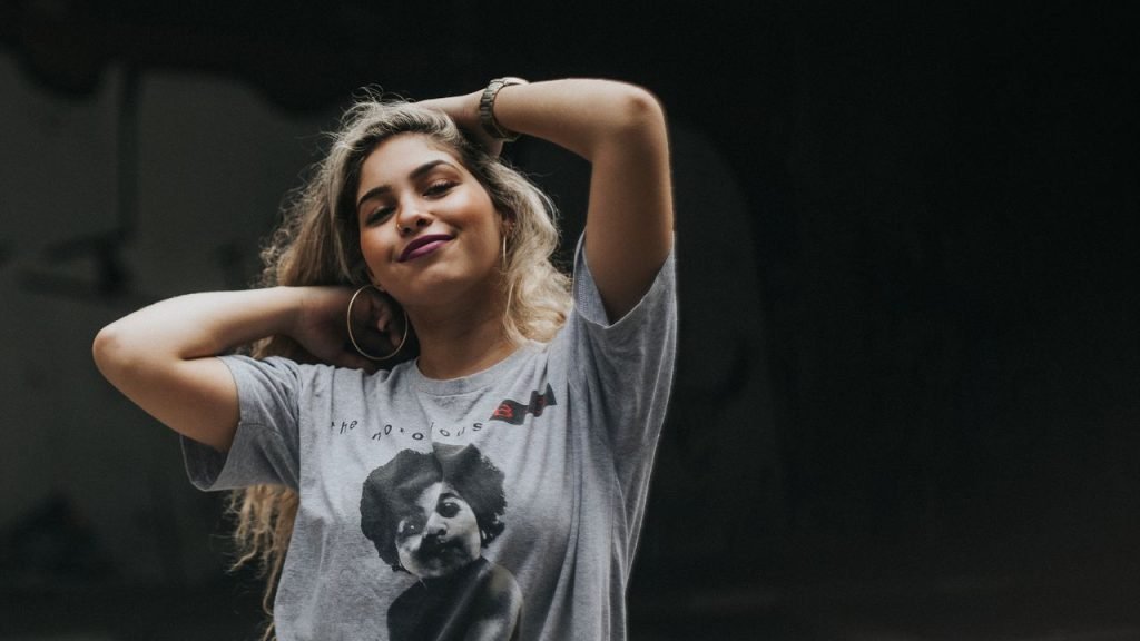 Young woman with blonde hair posing confidently in a The Notorious B.I.G. t-shirt against a dark background.